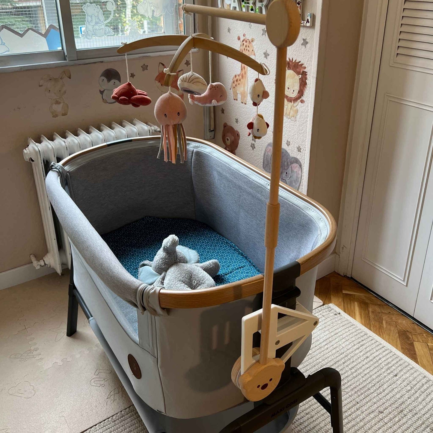 Full view on Maxi-cosi_Iora white CribClamp attached to the bedside crib with a wooden musical mobile. View of the Maxi-cosi co-sleeper and baby mobile attachment adapter bracket from the head side of the bedside cot